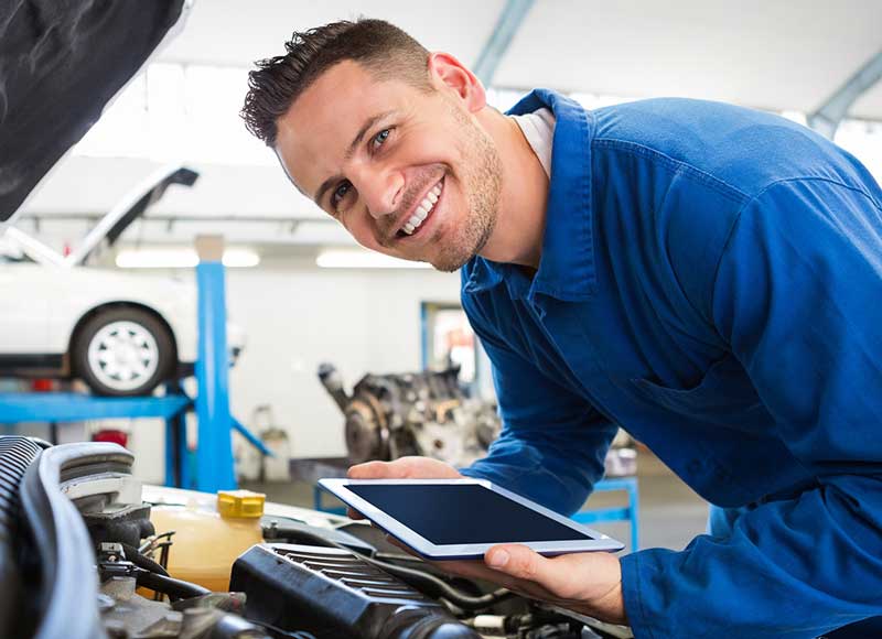 Smiling technician servicing a car engine while holding a tablet.